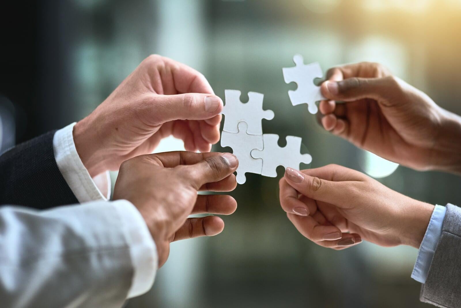 Three people are putting together their pieces of puzzle in the air. A fourth person is joining to complete the puzzle.