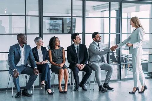 Woman greets the first candidate for a job interview. There are other candidates in smart professional outfits.