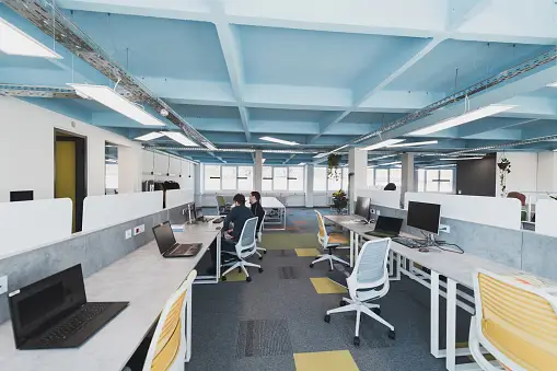 An office with people working at white desks. Computer setups.